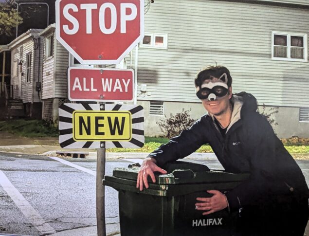 Stephen MacKay wearing a raccoon mask standing over and embracing a Halifax green bin next to a stop sign that has a "new" sign below it.